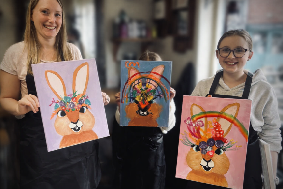 Two women holding paintings of rabbits with decorative headbands and colourful backgrounds.