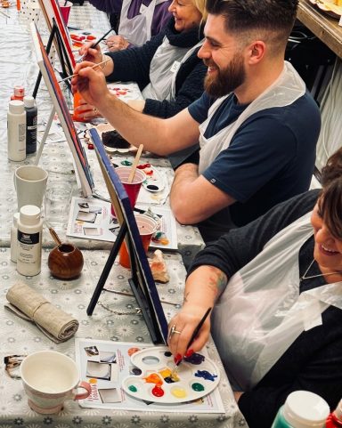 People painting on easels in a lively art class setting.