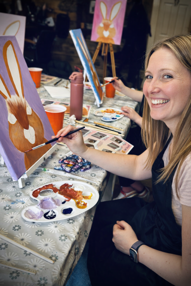 A smiling woman paints a rabbit on canvas at a creative workshop.