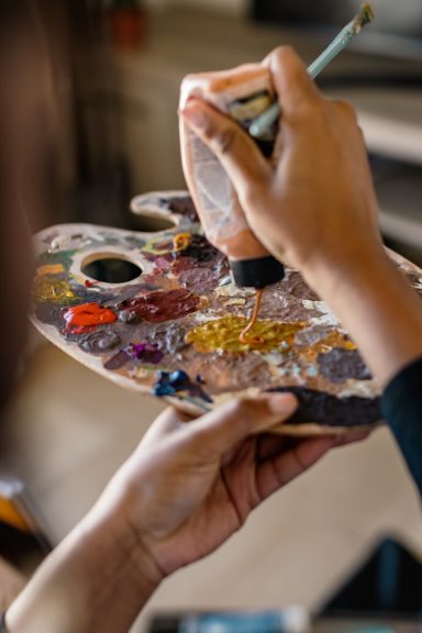 A person's hand holding a paintbrush over a palette filled with colourful paints.